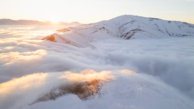 Snowy mountain range bathed in warm sunrise light above sea of clouds, aerial winter landscape