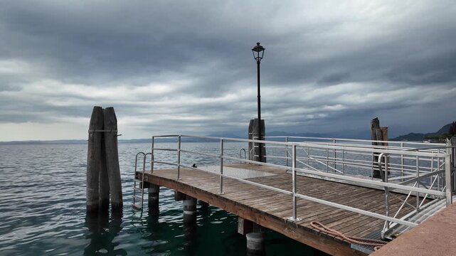 Bardolino, Italy - May 14, 2026 - Wooden pier with metal railings extends into calm lake water under cloudy sky