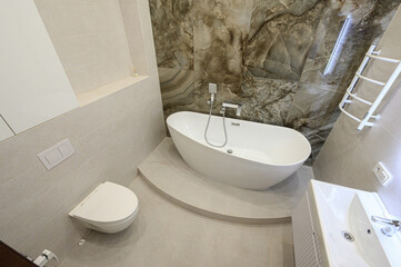 Fototapeta premium Modern bathroom with freestanding white bathtub on a raised platform, marble-effect wall tiles, concealed toilet, white sink, and chrome fixtures