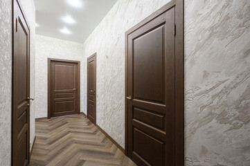 Fototapeta premium Narrow hallway with dark wooden panel doors, herringbone wood flooring, textured beige walls, and recessed ceiling lights creating a warm, modern atmosphere