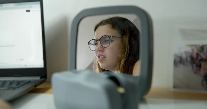 Young Woman Looking in Mirror: A young woman with glasses looks intently into a mirror, her reflection showing concern or deep thought. The scene is set at a desk with a laptop and other items.