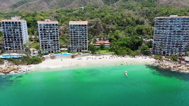 Aerial view of Las Gemelas Beach with people on the sand, high-rise resort buildings, and turquoise water against lush green hills in Puerto Vallarta, Jalisco, Mexico.