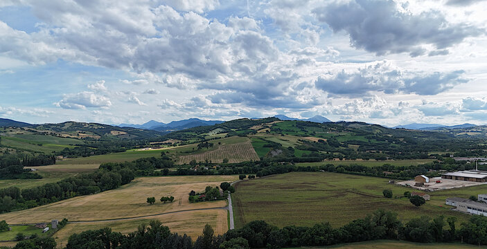 Panoramica di colline coltivate e campi verdi alternati a zone boschive, con strada rurale e strutture agricole sullo sfondo, sotto cielo parzialmente nuvoloso. Atmosfera ampia, naturale e luminosa
