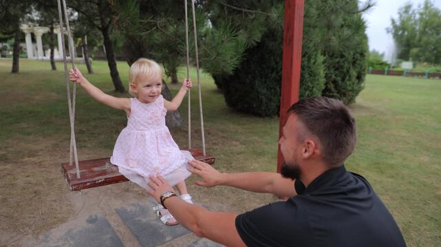 Young caring father pushing his joyful toddler daughter on a swing at a green park playground, creating a beautiful family memory during a warm summer day with lots of positive emotions