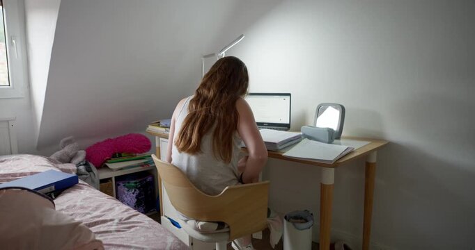 Student Studying at Desk: A young student with long brown hair sits at a wooden desk, focused on her laptop and books in a cozy room, illuminated by a desk lamp.