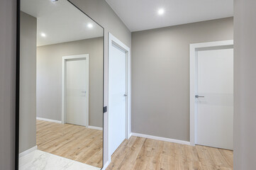 Fototapeta premium modern hallway with light grey walls, light wooden flooring, white doors, a large floor-length mirror, and recessed ceiling lighting