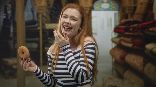 Redhead woman in striped off shoulder top holding a glazed donut, hand to mouth laughing while standing among stacked rugs in a building carpet shop; joyful laughter.