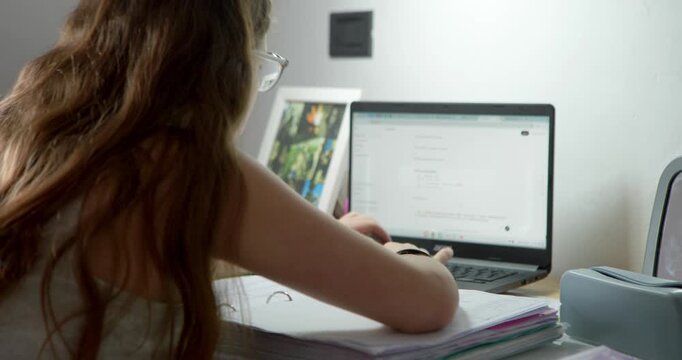 Girl Learning Online: A young girl with long, brown hair wearing glasses is focused on her laptop, engrossed in her online studies. A desk is filled with books and papers.