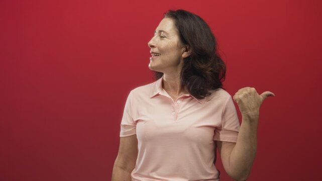 Woman middle aged smiles looking right and points thumb backward while wearing pink polo against red backdrop in studio; confidence.