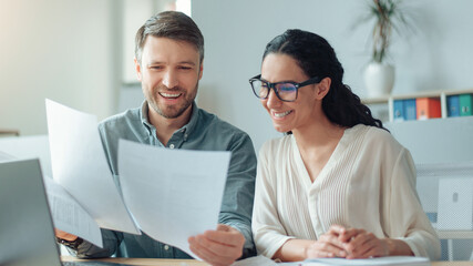 Smiling Caucasian Man And Woman Analyzing Documents And Discussing Ideas In A Bright Modern Office, Collaborating And Sharing Insights As Part Of Their Professional Work Environment. Business Concept © Prostock-studio