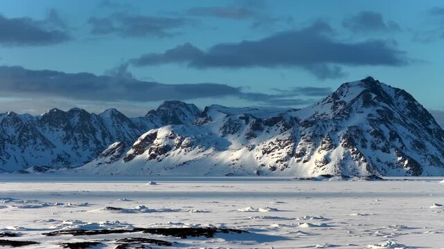 Aerial view of snow-covered mountain peaks and a vast frozen ice field under a cloudy blue sky in Tasiilaq, Sermersooq Municipality, Greenland.
