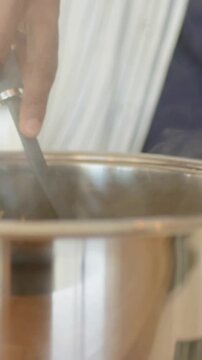 Vertical video: Stirring navy-sleeved hand with gray ladle at stove steam making tongs gripping pot