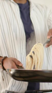 Vertical video: Lifting pasta from pot with tongs, cook in striped top guiding into pan finishing