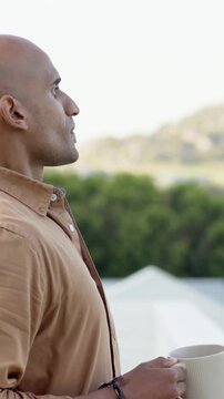 Vertical video: Pulling back camera, man holding mug on balcony, tan shirt, gazing, copy space