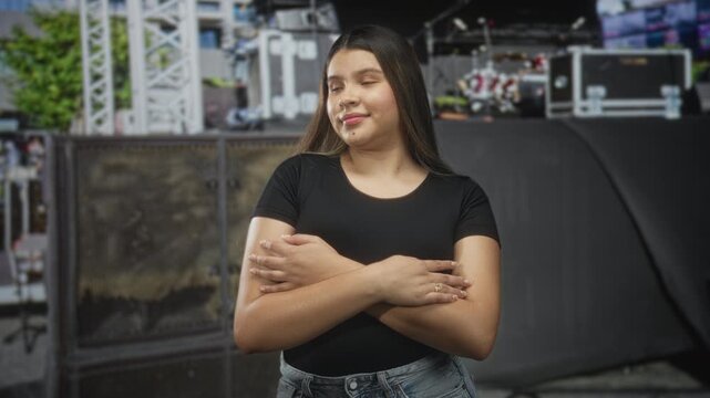 Young woman with arms crossed across chest on concert stage beside pa speakers and flight cases, wearing black top and jeans; quiet confidence.