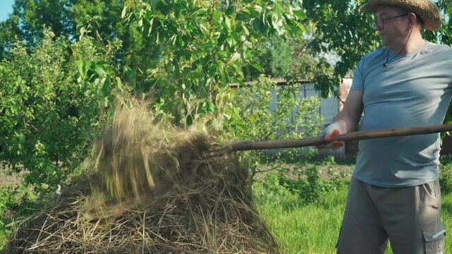 Gardener in straw hat rakes hay from a pile in a sunny garden surrounded by trees and greenery, showcasing the process of yard maintenance and care