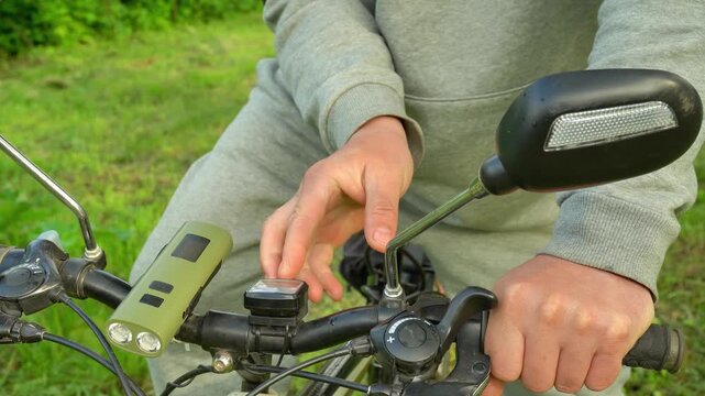 Person in gray outfit adjusts bicycle light on handlebars while preparing for a ride in a green outdoor setting with grass and trees in the background