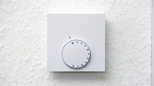 A white wall features a light switch, electrical outlet, and heating control panel arranged in a clean, minimal interior setup. The simple composition highlights everyday home fixtures on a smooth whi