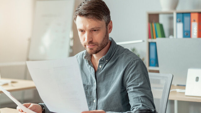 Focused Caucasian Male Professional In His 30s Reading Documents At Modern Office Workspace, Sitting At Desk With Laptop And Papers, Engaged In Work. Business Concept