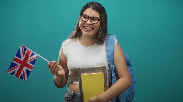 Young woman holding uk flag and yellow books, smiling with hand visible and backpack strap, studio; study abroad pride.