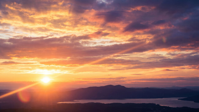 Heavenly sunset panorama, expansive sky filled with luminous cloud layers glowing in gold, coral, and purple hues, bright sun positioned off-center, cinematic lens flare, soft dark horizon below, phot