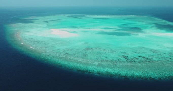 Aerial view of a vast coral reef atoll with turquoise waters surrounded by the deep blue Indian Ocean under a clear sky in Dhidhdhoo, Haa Alif, Maldives.