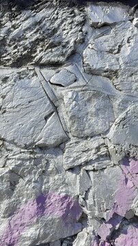 Close-up vertical shot of an industrial stone wall coated in metallic silver paint with purple accents, highlighting jagged edges and deep rock crevices.