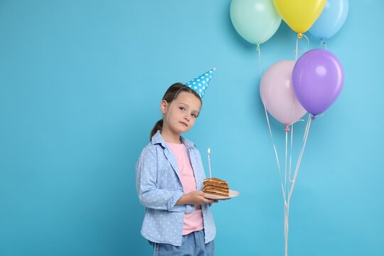 Little girl holding piece of cake with burning candle and colorful balloons on light blue background