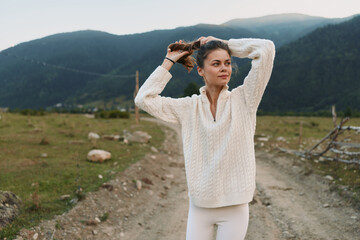 Fototapeta premium Young woman wearing a cream knit sweater stands on a dirt mountain road, adjusting her hair with rugged hills in the background, conveying a relaxed outdoor lifestyle.
