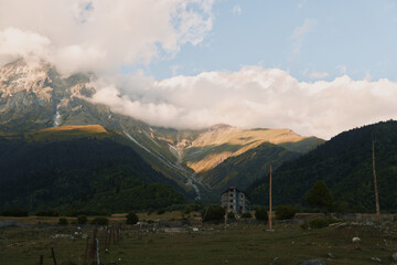 Naklejka premium Quiet mountain valley scene shows a small weathered house in a grassy field beneath dramatic clouds with distant peaks forming a rugged backdrop and a sense of solitude.