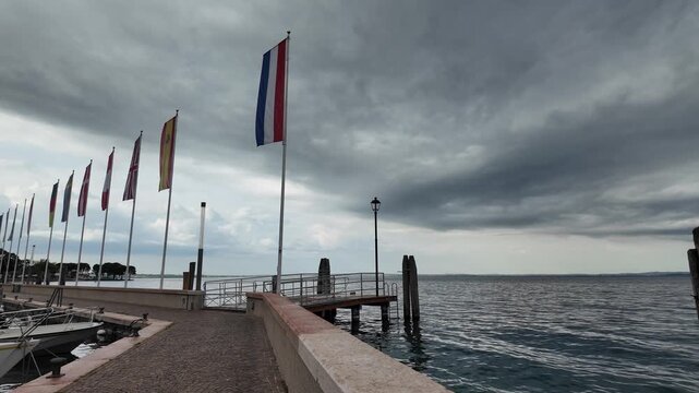 Bardolino, Italy - May 14, 2026 - Row of flags on a pier overlooking a calm lake under a cloudy sky