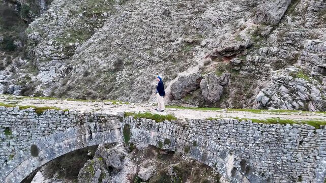 Aerial tracking shot pulling back from host Charles Huang standing on the historic stone arches of the Ali Pasha Bridge aqueduct in Gjirokaster, Albania