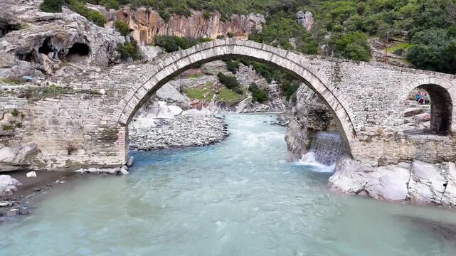 Push-in camera movement flying straight toward and under the historic stone arch of the Kadiut Bridge (Ura e Kadiut) over the rushing Lengarica River in Permet, Albania