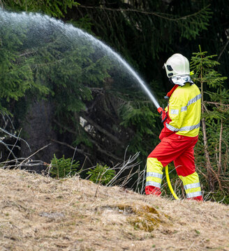 Firefighter Suppressing Wildfire at Forest Edge in Switzerland, Firefighter in protective gear using a water hose to suppress a wildfire or vegetation fire at the edge of a forest in Switzerland. Emer