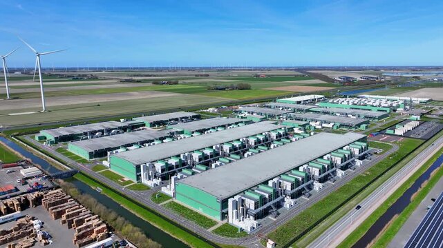 Aerial view of a modern industrial complex with green buildings and a wind turbine under a clear blue sky in Middenmeer, North Holland, Netherlands.