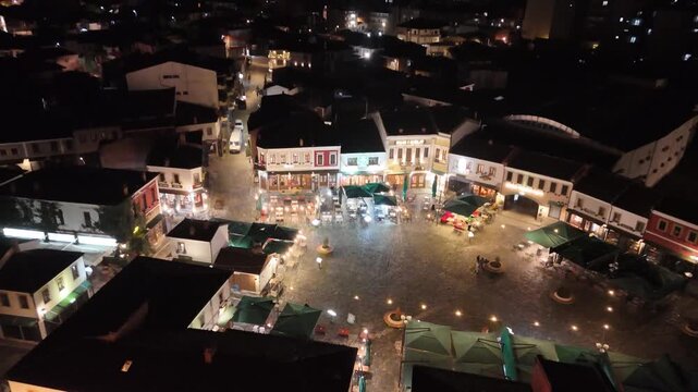 Aerial tracking shot moving forward over illuminated traditional facades, cafes, and outdoor restaurant patios in the main plaza of the historic Korca Old Bazaar at night