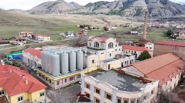Aerial tracking shot over the historic industrial facades, modern steel fermentation silos, and prominent brick chimney of the Birra Korca factory in Albania