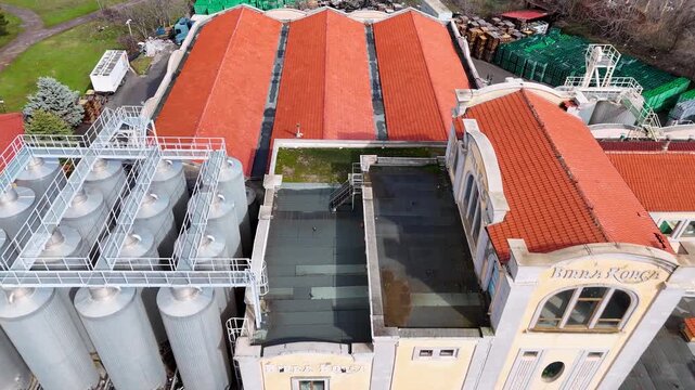Aerial tracking shot over the historic industrial facades, modern steel fermentation silos, and prominent brick chimney of the Birra Korca factory in Albania