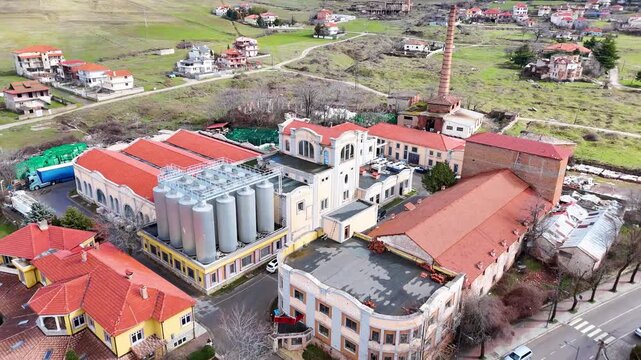Aerial tracking shot pulling back over the historic industrial facades, modern steel fermentation silos, and prominent brick chimney of the Birra Korca factory in Albania