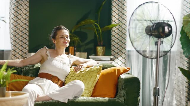 A serene woman on a green velvet sofa, with eyes closed and a gentle smile, enjoys the cooling breeze from a standing fan in a sunlit living room, capturing tranquil heat relief.