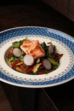 Close-up of a gourmet salad featuring grilled salmon fillets, fresh arugula, radish slices, and bell peppers served on an ornate blue patterned plate. Elegant restaurant seafood dish