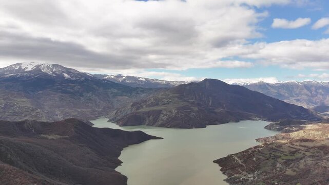 Aerial tracking shot pulling back over the vast, pale-green waters of the Charvak Reservoir lake, framing the massive rolling hills and distant snow-capped mountain ranges under an overcast sky