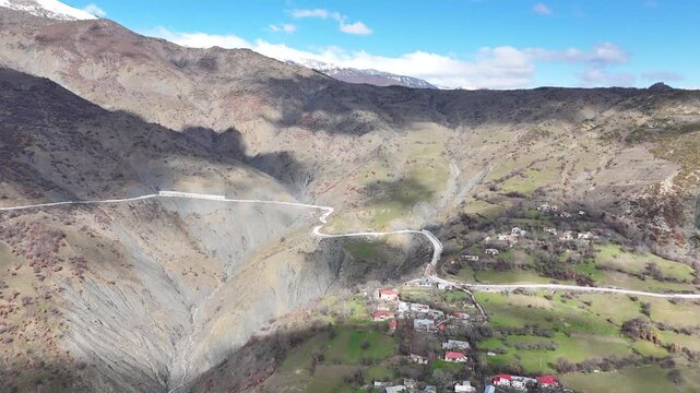 Aerial tracking shot panning leftward over a winding mountain highway and a small hillside village nestled below rugged alpine slopes under an overcast blue sky