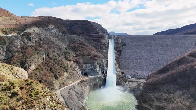 Aerial tracking shot moving forward toward the concrete spillway of Moglic&euml; Dam, capturing a powerful rush of cascading water splashing into the plunge pool below the massive rockfill wall