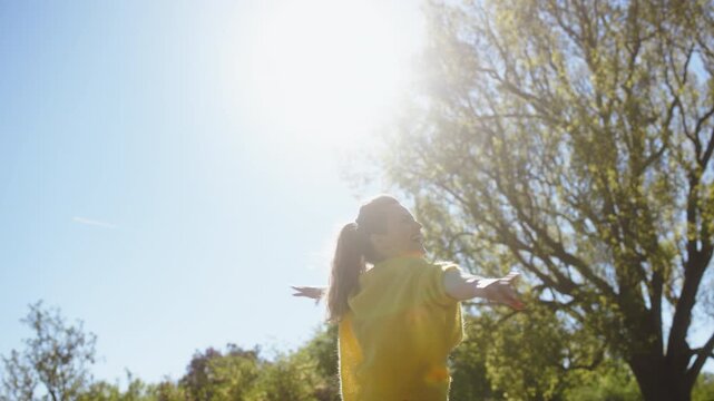 A woman in a yellow sweater spins with joy in a sun-drenched park, expressing freedom and happiness through movement during a beautiful spring day in nature.