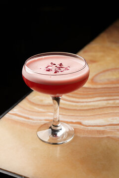 Close-up of a red alcoholic cocktail with pink foam layer, garnished with dry petals in a coupe glass on a textured bar counter. Elegant nightlife and mixology concept. Dark background.