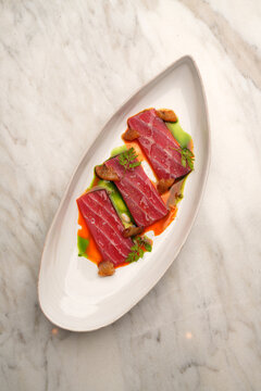 Top view of elegant tuna tataki slices served with citrus wedges, green herb oil, and ponzu sauce on an oval plate over a marble table. Gourmet seafood appetizer in a fine dining restaurant.