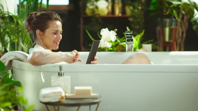 A middle-aged woman enjoys a luxurious bubble bath, leisurely scrolling on her tablet amidst lush greenery, blending modern connectivity with tranquil self-care.