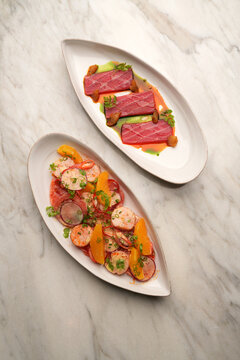 Top view of two elegant seafood dishes, tuna tataki sashimi with sauce and grilled scallop salad with oranges, served on oval plates over a light marble background. High-end restaurant catering