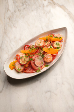 High angle view of a gourmet salad with grilled scallops, orange segments, radish slices, and tomatoes served on an oval ceramic plate over a marble table. Elegant restaurant seafood appetizer